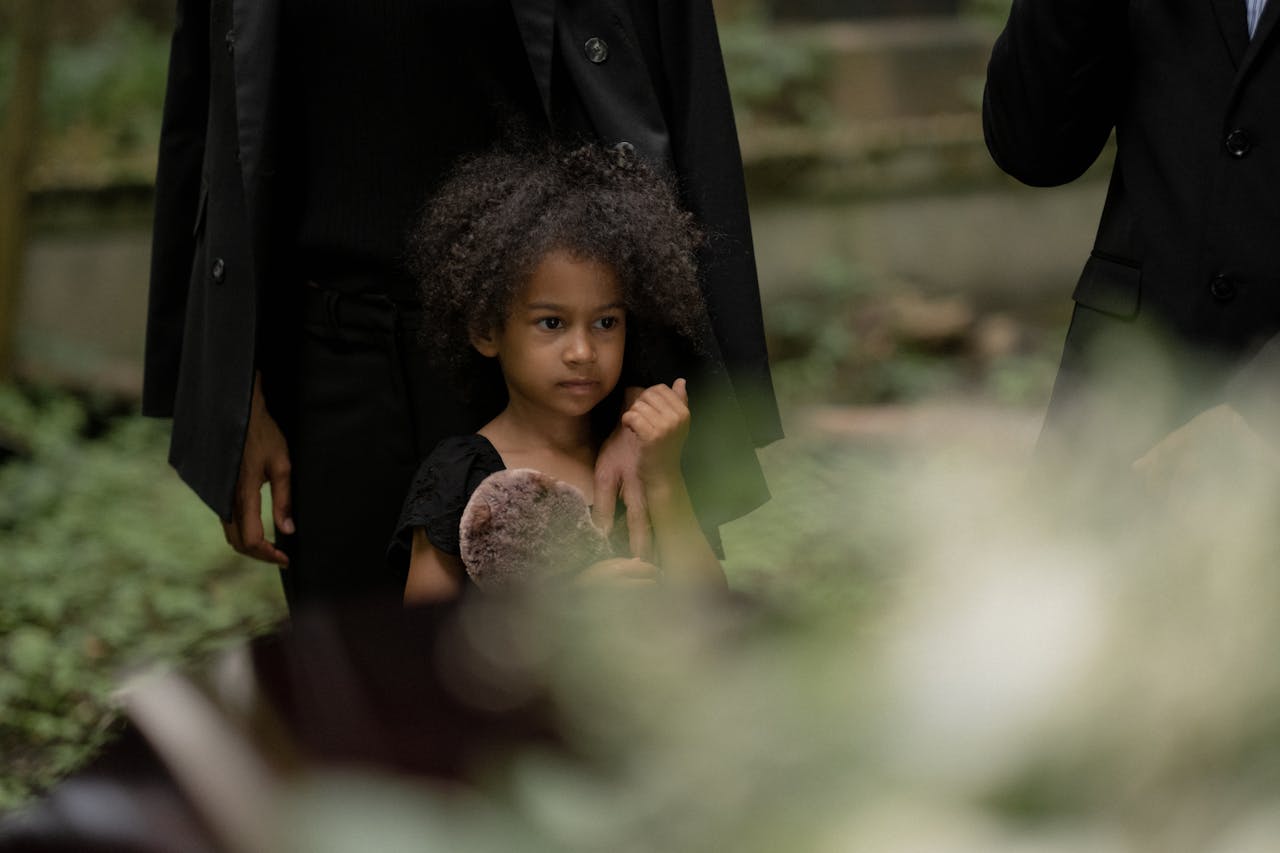 A young girl holding a stuffed toy during an outdoor funeral, conveying sadness.